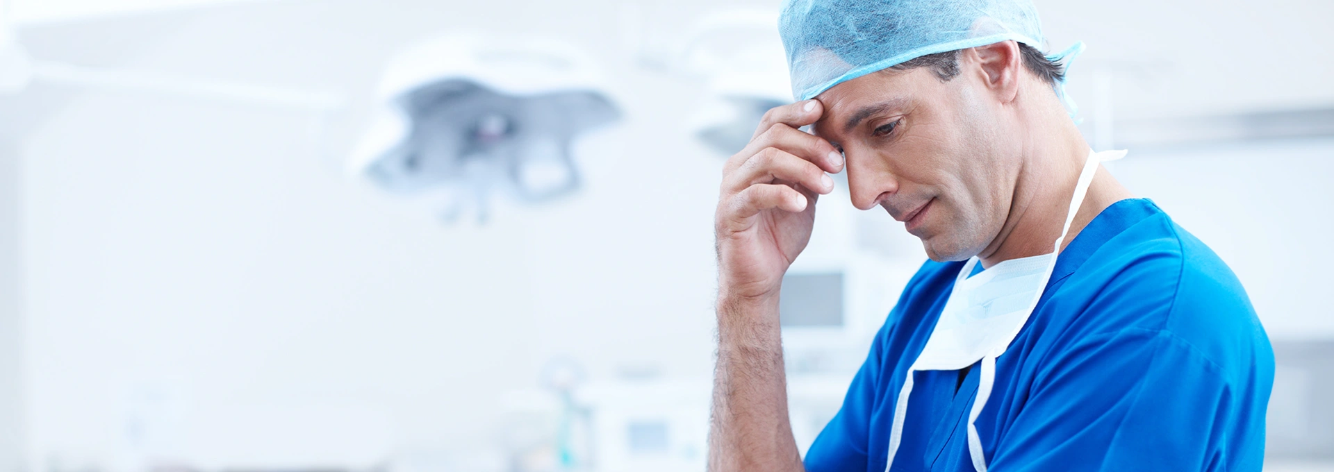 A male medical professional in blue scrubs and a surgical cap appears thoughtful and concerned in a clinical setting.