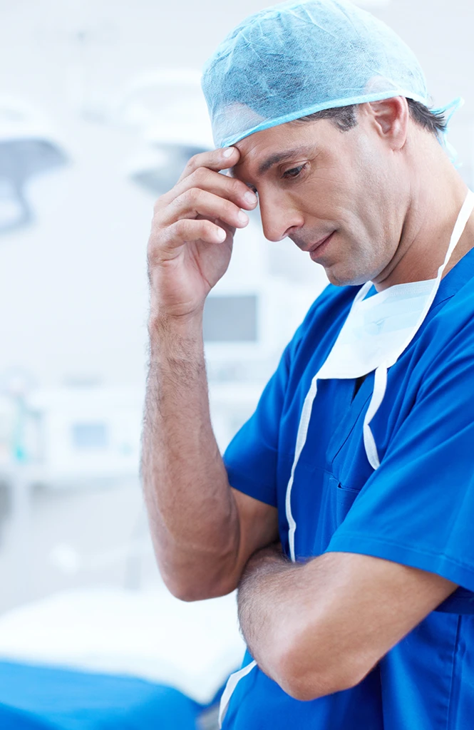 A male medical professional in blue scrubs and a surgical cap appears thoughtful and concerned in a clinical setting.